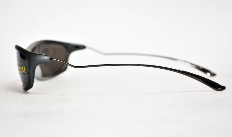 May include: A pair of black sunglasses with a thin metal frame and dark gray lenses. The sunglasses have a CE marking on the side.