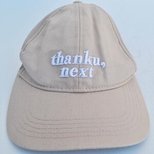 May include: A beige baseball cap with white embroidered text that reads "thanku, next".
