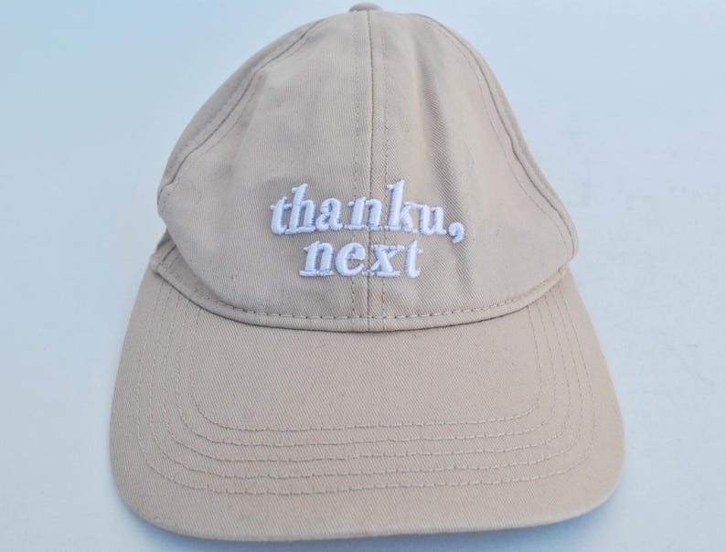 May include: A beige baseball cap with white embroidered text that reads "thanku, next".