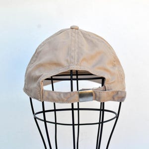 May include: A beige baseball cap with a black adjustable strap and a silver buckle. The cap is on a black metal display stand.