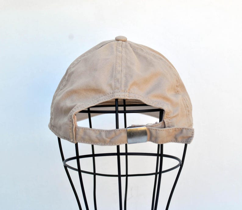 May include: A beige baseball cap with a black adjustable strap and a silver buckle. The cap is on a black metal display stand.