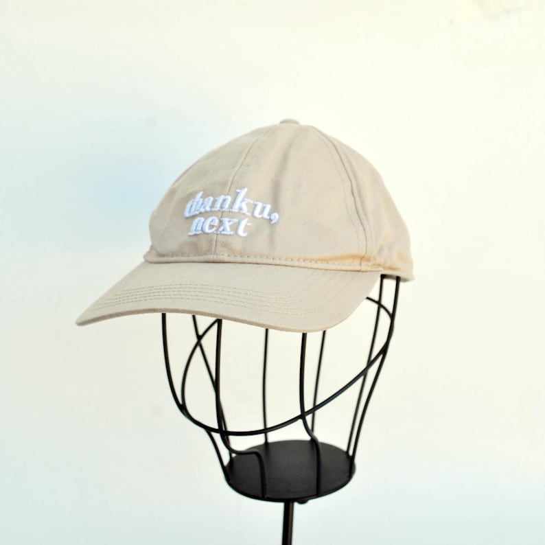 May include: A beige baseball cap with white embroidered text that reads "thanku, next".