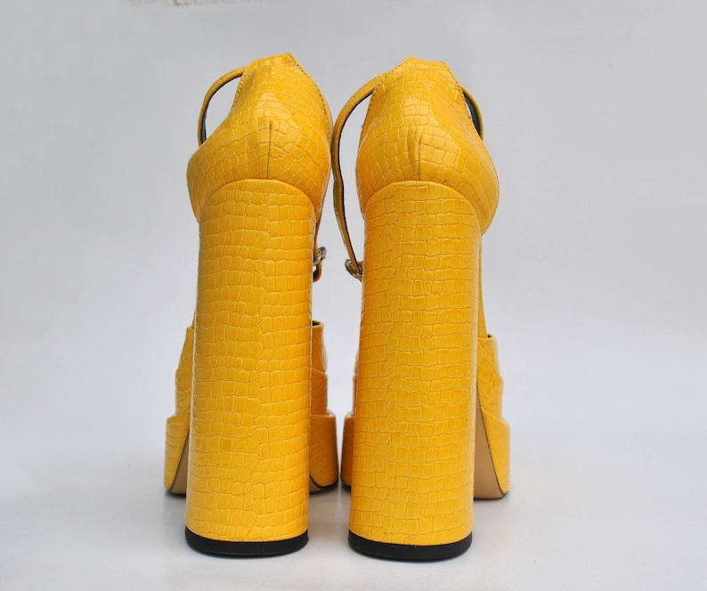 May include: A pair of yellow platform sandals with a crocodile-embossed texture. The sandals have a thick, chunky heel and an ankle strap.