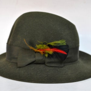 May include: A dark green felt fedora hat with a wide brim and a black ribbon. The hat is decorated with a small, colorful feather accent.