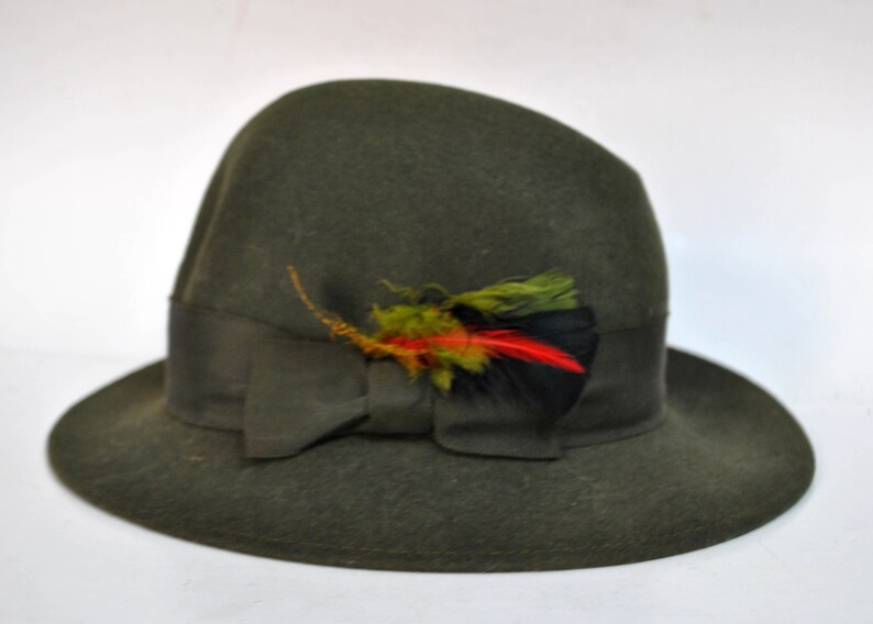 May include: A dark green felt fedora hat with a wide brim and a black ribbon. The hat is decorated with a small, colorful feather accent.