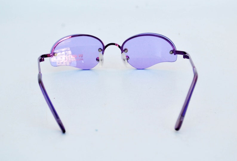 May include: Purple rimless sunglasses with oval lenses and a thin metal frame. The lenses are a light purple colour, and the frame is a darker purple. The sunglasses have a modern, minimalist design.