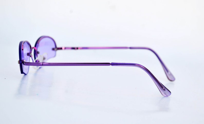 May include: A pair of purple-tinted, round-lens spectacles with a slender, metallic frame. The glasses feature a minimalist design, with a bridge and temples in a matching purple shade. The lenses are a solid purple colour, and the overall aesthetic is sleek and modern.