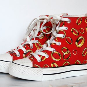 K&ouml;nnte beinhalten: Rote High-Top-Sneaker mit wei&szlig;er Schn&uuml;rung und wei&szlig;er Gummisohle. Die Sneaker sind mit einem Muster aus goldenen Ringen verziert.