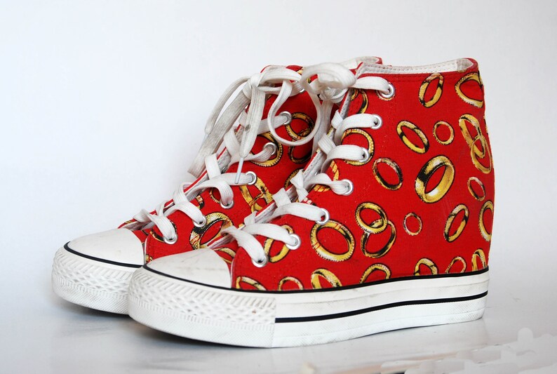 K&ouml;nnte beinhalten: Rote High-Top-Sneaker mit wei&szlig;er Schn&uuml;rung und wei&szlig;er Gummisohle. Die Sneaker sind mit einem Muster aus goldenen Ringen verziert.