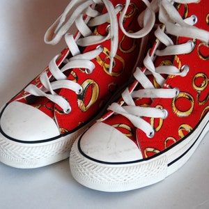 K&ouml;nnte beinhalten: Ein Paar rote High-Top-Sneaker mit wei&szlig;en Schn&uuml;rsenkeln und einer wei&szlig;en Gummisohle. Die Sneaker sind mit einem Muster aus goldenen Ringen bedeckt.