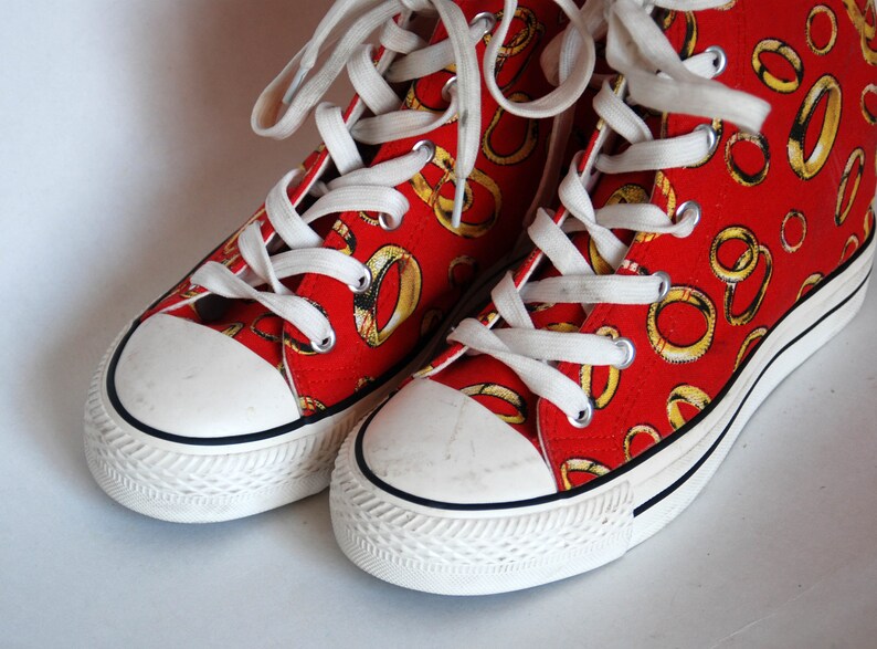 K&ouml;nnte beinhalten: Ein Paar rote High-Top-Sneaker mit wei&szlig;en Schn&uuml;rsenkeln und einer wei&szlig;en Gummisohle. Die Sneaker sind mit einem Muster aus goldenen Ringen bedeckt.