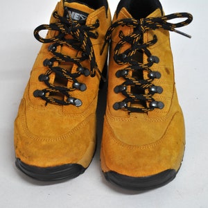 May include: A pair of yellow suede hiking boots with black laces. The boots have a black rubber sole.