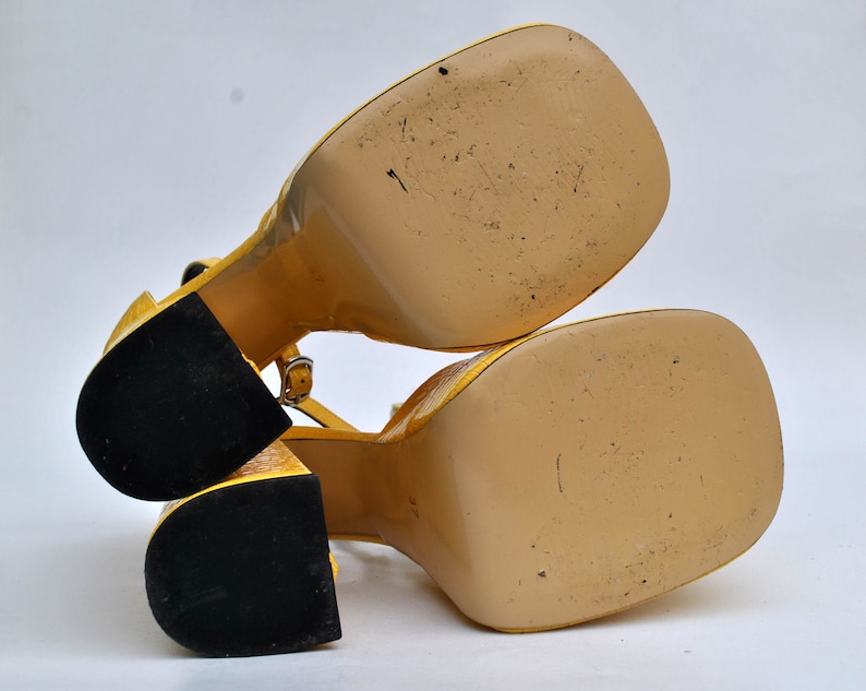 May include: A pair of yellow platform sandals with black soles. The sandals have a strap across the ankle and a buckle fastening.