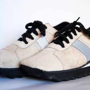May include: A pair of white furry trainers with black laces and a silver stripe on each side. The trainers have a black rubber sole.