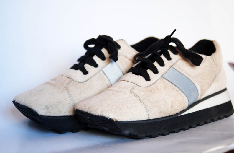 May include: A pair of white furry trainers with black laces and a silver stripe on each side. The trainers have a black rubber sole.