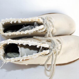 May include: A pair of white canvas boots with frayed edges and laces. The boots are tied together and are lying on a white surface.