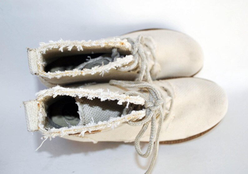 May include: A pair of white canvas boots with frayed edges and laces. The boots are tied together and are lying on a white surface.