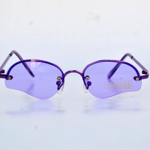 May include: Rimless sunglasses in purple with oval lenses. The frames and arms are purple. The lenses have a distinctive shape and a UV 400 sticker. The glasses have a CE mark.
