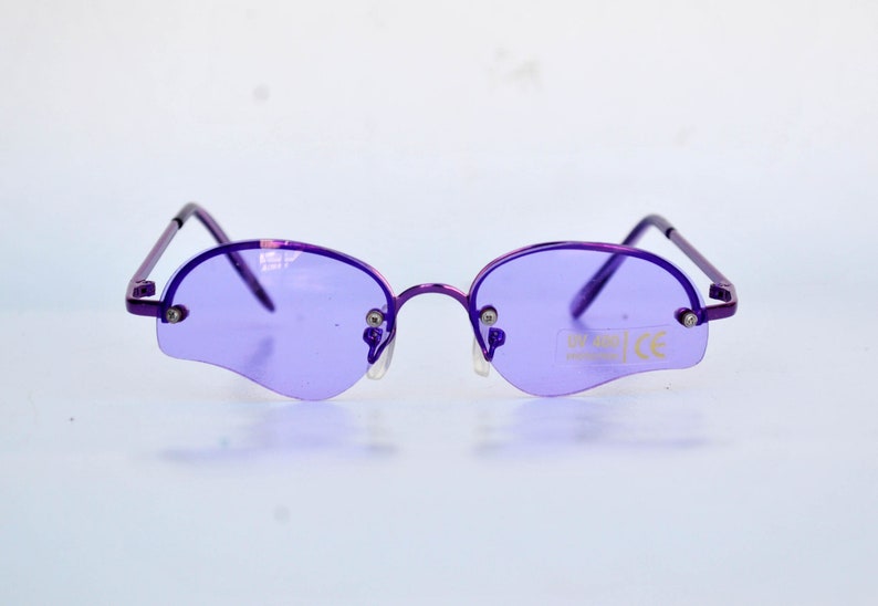 May include: Rimless sunglasses in purple with oval lenses. The frames and arms are purple. The lenses have a distinctive shape and a UV 400 sticker. The glasses have a CE mark.