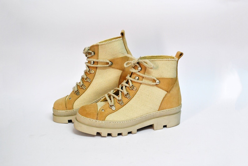 May include: A pair of beige canvas and suede hiking boots with laces. The boots have a chunky white sole.