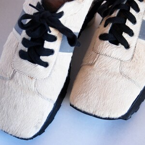 May include: A pair of white furry trainers with black laces and a silver reflective stripe on each side.