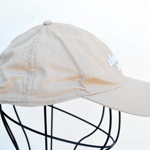 May include: A beige baseball cap with a white embroidered logo that says "The Daily Grind".
