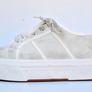 May include: White platform sneakers with a white lace-up closure. The shoes have a thick white sole with a brown rubber trim.