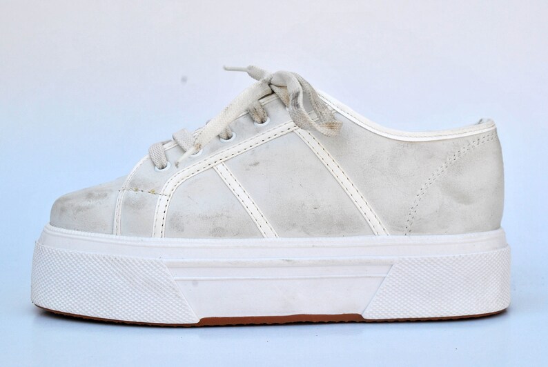 May include: White platform sneakers with a white lace-up closure. The shoes have a thick white sole with a brown rubber trim.