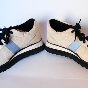 May include: A pair of white calf hair trainers with black laces and a blue stripe on each side. The trainers have a black rubber sole.