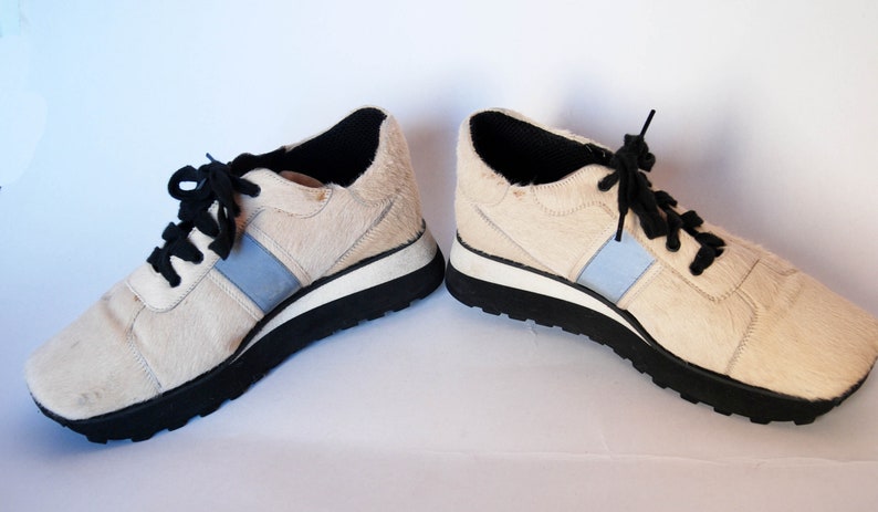 May include: A pair of white calf hair trainers with black laces and a blue stripe on each side. The trainers have a black rubber sole.