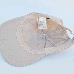 May include: A beige baseball cap with a label that reads "Made in Bangladesh, 100% Cotton, Size: One Size", and a small silver buckle on the back.