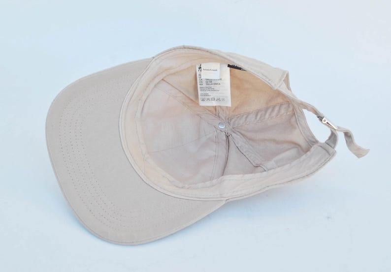 May include: A beige baseball cap with a label that reads "Made in Bangladesh, 100% Cotton, Size: One Size", and a small silver buckle on the back.