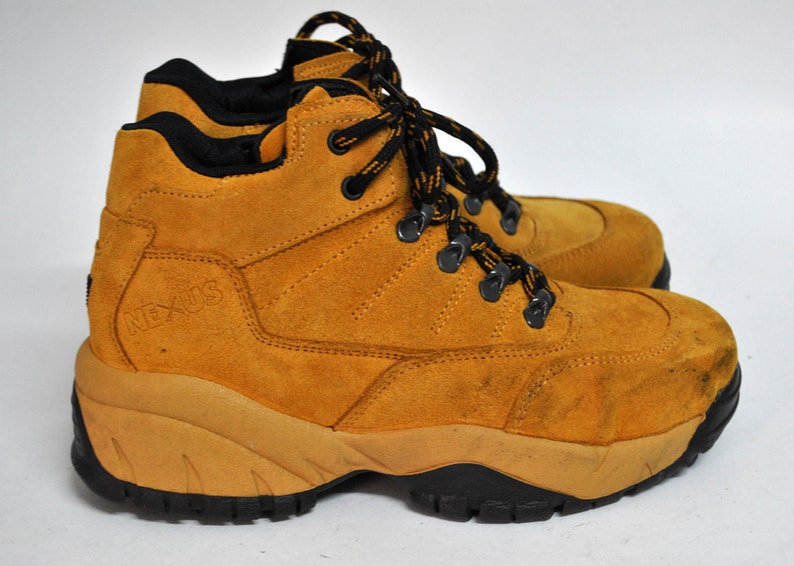 May include: Tan suede hiking boots with black laces and a black "NEXUS" logo on the side. The boots have a thick, rugged sole.