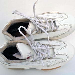 Puede incluir: Un par de zapatillas blancas de cuero con cordones blancos. Las zapatillas tienen un dise&ntilde;o texturizado y la palabra "SKECHERS" es visible en el interior de cada zapato.
