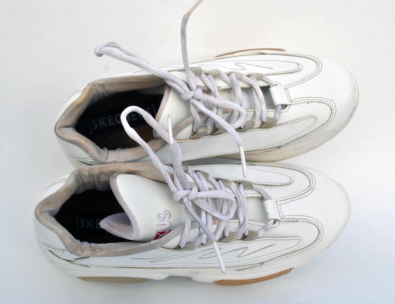 Puede incluir: Un par de zapatillas blancas de cuero con cordones blancos. Las zapatillas tienen un dise&ntilde;o texturizado y la palabra "SKECHERS" es visible en el interior de cada zapato.