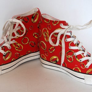 K&ouml;nnte beinhalten: Rote High-Top-Canvas-Sneaker mit wei&szlig;er Sohle und wei&szlig;en Schn&uuml;rsenkeln. Die Sneaker sind mit einem Muster aus goldenen Ringen bedeckt.