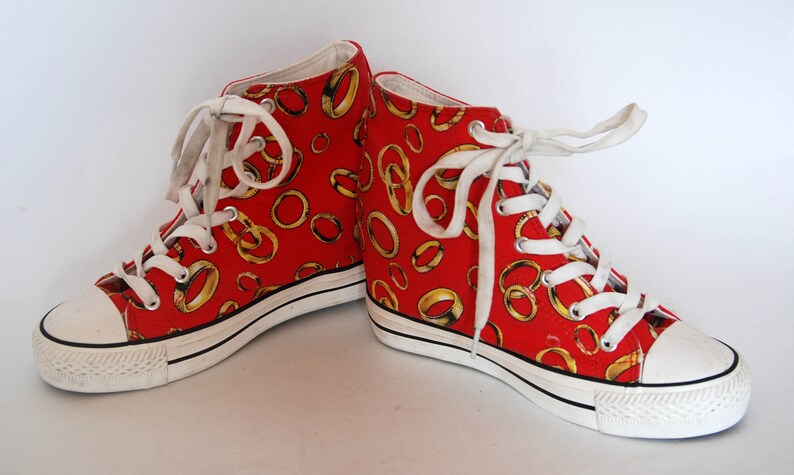 K&ouml;nnte beinhalten: Rote High-Top-Canvas-Sneaker mit wei&szlig;er Sohle und wei&szlig;en Schn&uuml;rsenkeln. Die Sneaker sind mit einem Muster aus goldenen Ringen bedeckt.