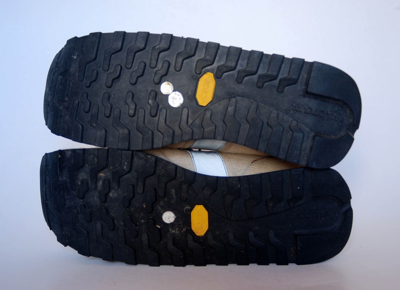 May include: Close-up of the bottom of a pair of beige trainers with black soles. The soles have a textured pattern and yellow accents.