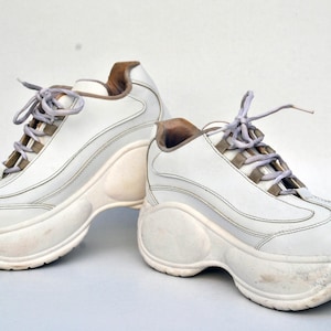 Puede incluir: Un par de zapatillas blancas de plataforma con suela gruesa. Las zapatillas tienen una parte superior de cuero blanco y un cierre de cordones blanco.