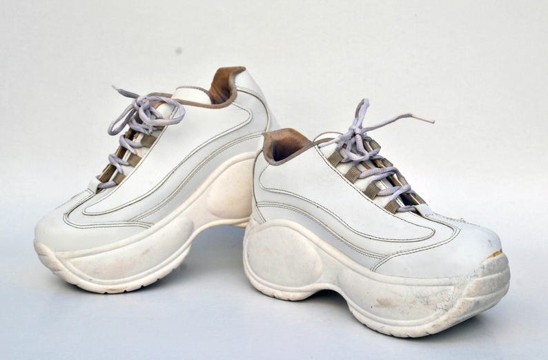 Puede incluir: Un par de zapatillas blancas de plataforma con suela gruesa. Las zapatillas tienen una parte superior de cuero blanco y un cierre de cordones blanco.