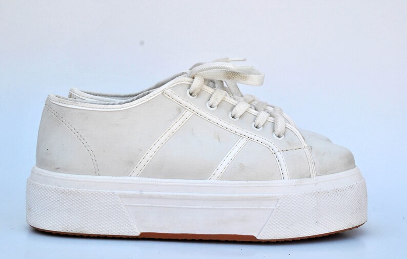 May include: White platform sneakers with a lace-up closure. The shoes have a thick white sole and a slightly distressed look.
