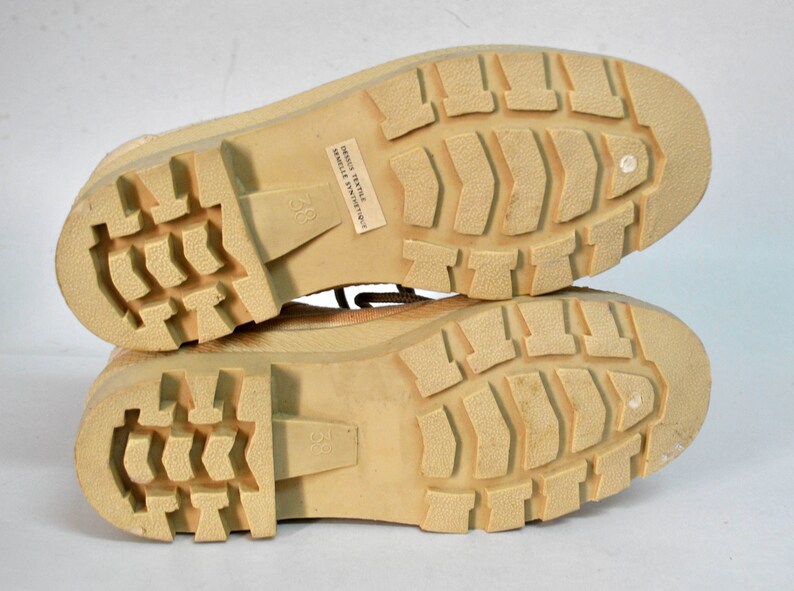 May include: Close-up of the soles of a pair of tan canvas boots. The boots have a textured rubber sole and the text "DESIGN TEXTILE SEMELLE SYNTHETIQUE" is printed on the sole of one boot.