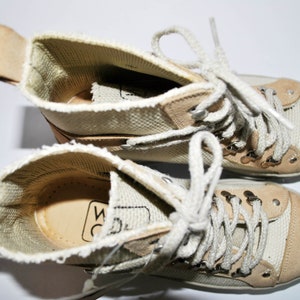 May include: A pair of beige canvas high-top sneakers with white laces and a brown leather trim. The sneakers have a woven texture and the brand name "WC" is visible on the inside of the shoe.