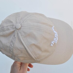 May include: A beige baseball cap with a white embroidered design that says "the days" on the side.