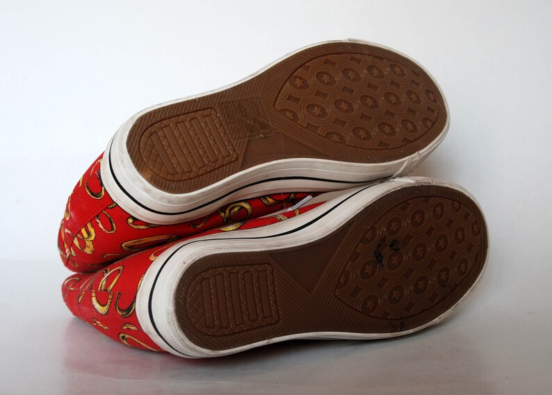 K&ouml;nnte beinhalten: Ein Paar rote Canvas-High-Top-Sneaker mit goldenem Muster. Die Schuhe haben wei&szlig;e Sohlen und braune Gummisohlen.