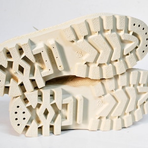 May include: Close-up of the white rubber soles of a pair of white sneakers. The soles have a textured tread pattern and multiple small holes.