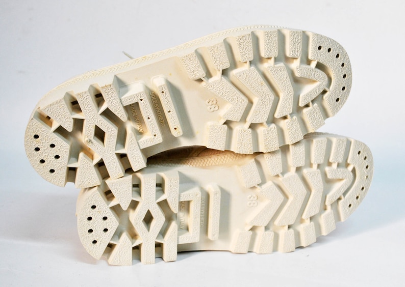May include: Close-up of the white rubber soles of a pair of white sneakers. The soles have a textured tread pattern and multiple small holes.
