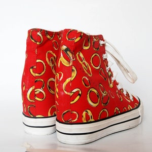 K&ouml;nnte beinhalten: Rote High-Top-Sneaker mit einem goldenen Ringmuster. Die Sneaker haben wei&szlig;e Schn&uuml;rsenkel und eine wei&szlig;e Sohle.