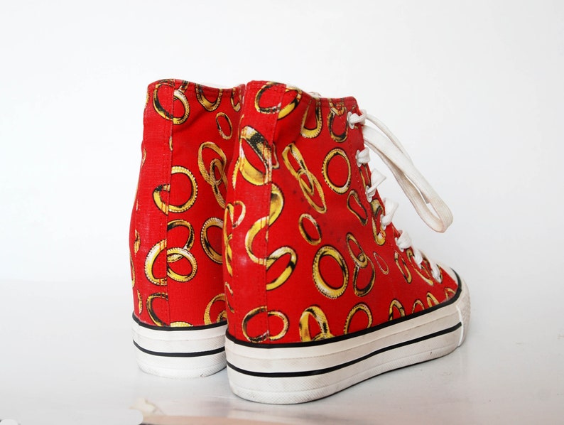 K&ouml;nnte beinhalten: Rote High-Top-Sneaker mit einem goldenen Ringmuster. Die Sneaker haben wei&szlig;e Schn&uuml;rsenkel und eine wei&szlig;e Sohle.
