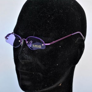 May include: Rimless sunglasses with purple lenses and a thin, purple metal frame. The oval lenses have a solid purple tint. The glasses are displayed on a black mannequin head. The lenses have the text "UV 400 PROTECTION" and "CE".